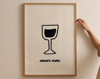 May include: A framed art print featuring a black and white illustration of a wine glass. The words "cheers mate." are printed in a black, sans-serif font below the glass. The frame is a light brown color.