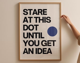 May include: A framed art print with the text "STARE AT THIS DOT UNTIL YOU GET AN IDEA" in bold black letters. A blue circle is to the right of the word "DOT". The frame is a light brown color.