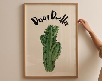 May include: A framed art print featuring a watercolor illustration of a green cactus with black spines. The words "Desert Dweller" are written in black script above the cactus. The frame is a light brown color.