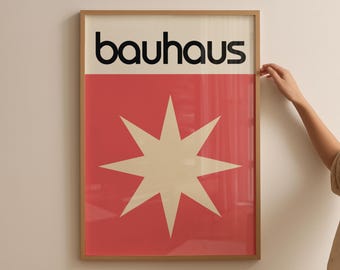 May include: Framed Bauhaus art print. The print features the word "bauhaus" in black letters above a red rectangle with a cream-colored starburst design. The frame is a light brown color.