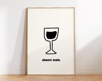 May include: A framed print featuring a simple black and white illustration of a wine glass filled with dark liquid. The words "cheers mate" are printed in a black, sans-serif font below the glass. The frame is a light wood color.