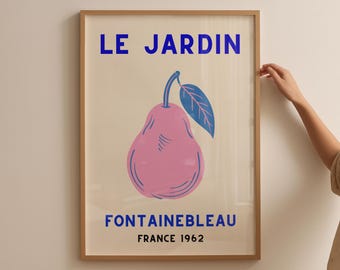 May include: A framed art print featuring a pink pear with a blue leaf, set against a cream background. The words "LE JARDIN" and "FONTAINEBLEAU FRANCE 1962" are printed in blue. The frame is a light brown color.