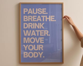 May include: A framed poster with a light blue background and the words "PAUSE. BREATHE. DRINK WATER. MOVE YOUR BODY." in a beige font. The frame is a light brown color. The poster is hanging on a white wall.