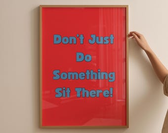 May include: A framed poster with a red background and blue text that reads "Don't Just Do Something Sit There!". The frame is a light brown color. The poster is hanging on a white wall.