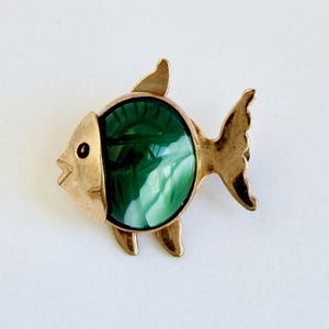 Puede incluir: Broche dorado con forma de pez, con una piedra ovalada de mármol verde y blanco en el centro. El broche tiene un diseño detallado con aletas y un pequeño ojo.