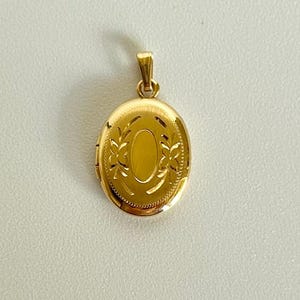 May include: A gold-colored oval locket pendant with a decorative leaf design. The pendant has a small loop at the top for a chain and a textured border. The locket is closed and sits on a white surface.