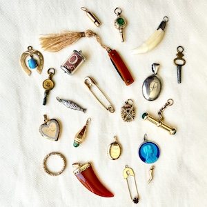 May include: A collection of assorted metal and stone charms and pendants, including a horseshoe with a blue stone, a small folding knife with a tassel, a silver fish, a heart-shaped locket, a red horn-shaped pendant, and various keys and safety pins.