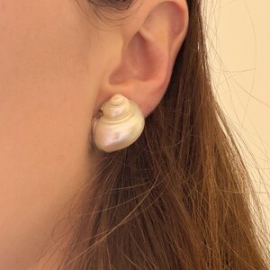 May include: A pair of pearl-colored seashell stud earrings. The earrings are round and have a spiral design, with a silver-colored metal backing. The earrings are worn on an ear.