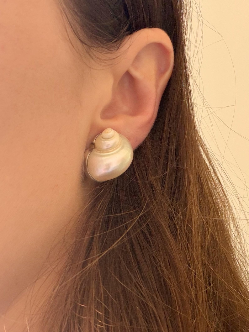 May include: A pair of pearl-colored seashell stud earrings. The earrings are round and have a spiral design, with a silver-colored metal backing. The earrings are worn on an ear.