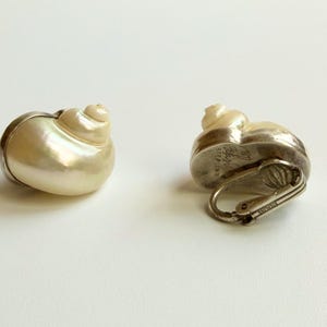 May include: Two seashell-shaped earrings. The earrings have a pearlescent, off-white shell design with a silver-colored metal base and clasp. One earring is lying on its side, and the other is upright.