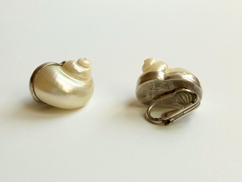 May include: Two seashell-shaped earrings. The earrings have a pearlescent, off-white shell design with a silver-colored metal base and clasp. One earring is lying on its side, and the other is upright.