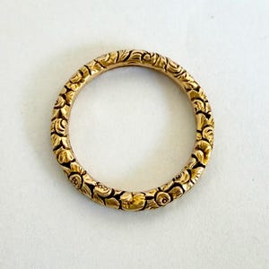 Könnte beinhalten: Ein goldfarbener Ring mit einem aufwendigen Blumenmuster. Der Ring ist kreisförmig und scheint aus Metall zu sein. Das florale Design bedeckt die gesamte Oberfläche des Rings und erzeugt ein strukturiertes Aussehen. Der Ring ist vor einem weißen Hintergrund platziert.