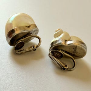 May include: A pair of vintage clip-on earrings. Each earring features a cream-colored, shell-shaped design with a silver-tone metal base and clip. The earrings are marked "STERLING".
