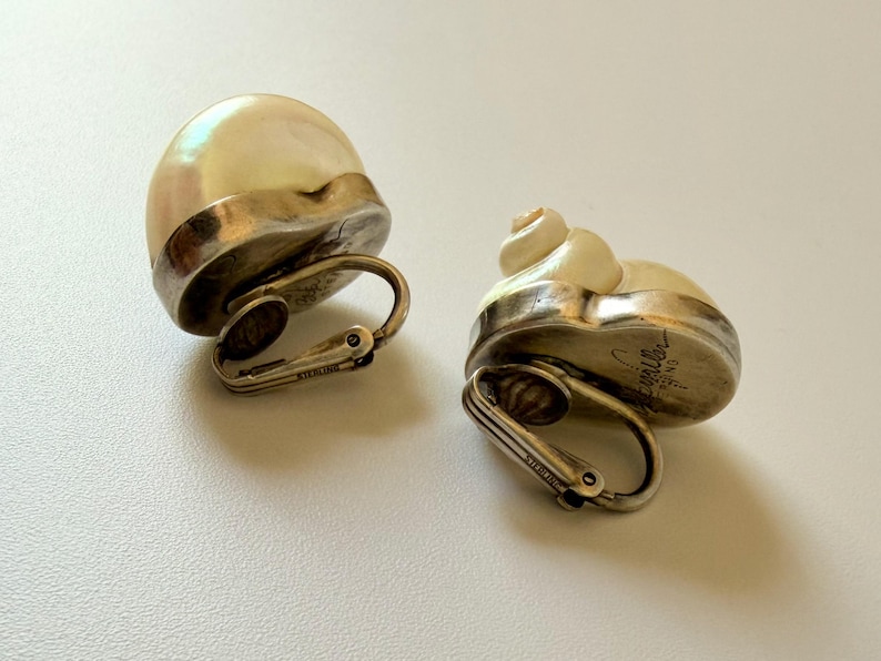 May include: A pair of vintage clip-on earrings. Each earring features a cream-colored, shell-shaped design with a silver-tone metal base and clip. The earrings are marked "STERLING".