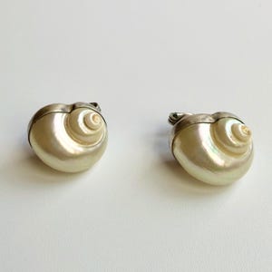 May include: Two decorative shell-shaped trinket boxes. The boxes are pearlescent white with a silver-colored metal trim. The spiral design of the shells is clearly visible. The boxes are on a white background.