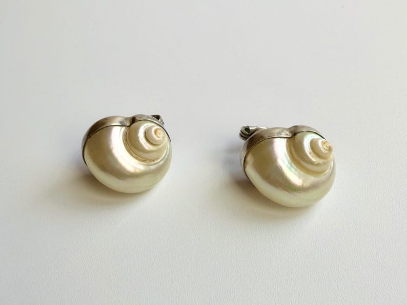 May include: Two decorative shell-shaped trinket boxes. The boxes are pearlescent white with a silver-colored metal trim. The spiral design of the shells is clearly visible. The boxes are on a white background.