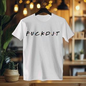 May include: White crew neck t-shirt with the text "FUCK DJ T" in a multi-colored font. The shirt is hanging on a black hanger against a blurred background.
