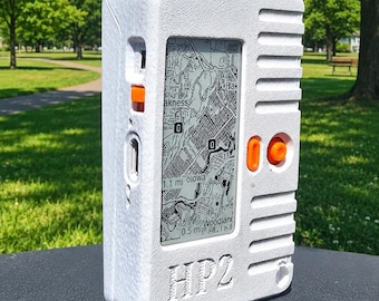 Peut inclure: Un appareil radio portable blanc à la finition texturée, doté d'une antenne noire, d'un écran vertical affichant une carte et de boutons orange. L'avant de l'appareil porte l'inscription HP2 en relief et comporte un interrupteur orange latéral.