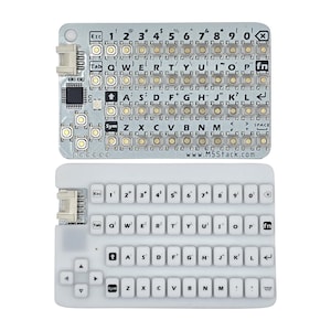 May include: Two electronic keyboard modules. The top module is silver with black keys and the text "www.M5Stack.com". The bottom module is white with white keys. Both modules have a variety of buttons and keys with letters, numbers, and symbols.