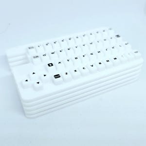 Könnte beinhalten: Eine weiße, kompakte Tastatur mit erhabenen, abgerundeten Tasten. Die Tasten sind mit schwarzen Buchstaben, Zahlen und Symbolen beschriftet. Die Tastatur hat ein geschichtetes Design und enthält Pfeiltasten und eine 'Sym'-Taste.