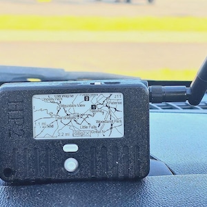 May include: A black rectangular electronic device with the text HP2 embossed on the side. It features a small screen displaying a map with location names like Totowa and Little Falls, a white button, and a black antenna extending from the right side.