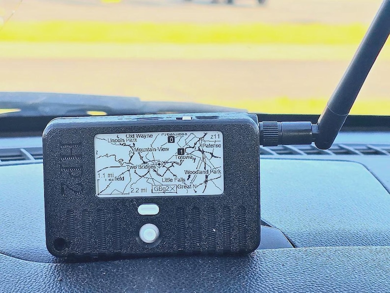May include: A black rectangular electronic device with the text HP2 embossed on the side. It features a small screen displaying a map with location names like Totowa and Little Falls, a white button, and a black antenna extending from the right side.