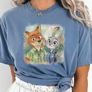 May include: Blue jean colored t-shirt featuring a colorful graphic of Nick Wilde and Judy Hopps from Zootopia. The image shows the characters smiling, with Nick in a green shirt and Judy in a police uniform. The text "BLUE JEAN" is at the bottom.