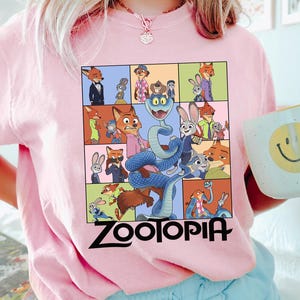 Puede incluir: Una camiseta rosa claro con un gr&aacute;fico colorido de personajes de la pel&iacute;cula animada "Zootopia". El dise&ntilde;o incluye una cuadr&iacute;cula de im&aacute;genes y la palabra "Zootopia" en negro. Se sostiene una taza blanca con una carita sonriente amarilla.