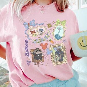 Puede incluir: Camiseta rosa claro con ilustraciones coloridas de personajes de la pel&iacute;cula "Zootr&oacute;polis". El dise&ntilde;o incluye retratos enmarcados, una guirnalda de luces y la palabra "Zootopia". Se ve una taza blanca con una carita sonriente amarilla.