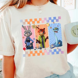 Puede incluir: Camiseta de color marfil con tres personajes de dibujos animados en marcos rectangulares con un patr&oacute;n de cuadros. Los personajes incluyen un conejo, un zorro y una serpiente. Una taza blanca con una carita sonriente se sostiene en la mano.