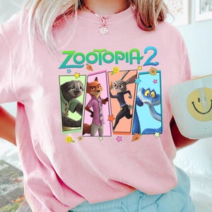 K&ouml;nnte beinhalten: Ein hellrosa T-Shirt mit dem Text "Zootopia 2" und einer Grafik von vier Charakteren aus dem Animationsfilm. Die Charaktere befinden sich in farbigen rechteckigen Rahmen. Eine wei&szlig;e Tasse mit einem gelben Smiley wird in der Hand gehalten.