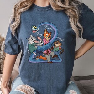 K&ouml;nnte beinhalten: Denimblaues T-Shirt mit einer Cartoon-Grafik von Filmfiguren. Die Grafik zeigt einen Fuchs, ein Kaninchen, einen Biber und eine Schlange. Die Namen der Charaktere sind dar&uuml;ber geschrieben. Das Wort "DENIM" ist unten rechts aufgedruckt.