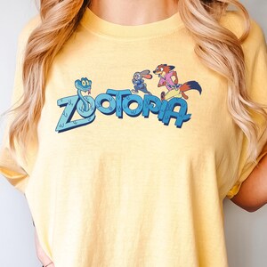 Op de afbeelding: Geel t-shirt met de tekst "Zootopia" in blauw, met stripfiguren. Het shirt heeft een ronde hals en korte mouwen. Het woord "BUTTER" staat onderaan het shirt.