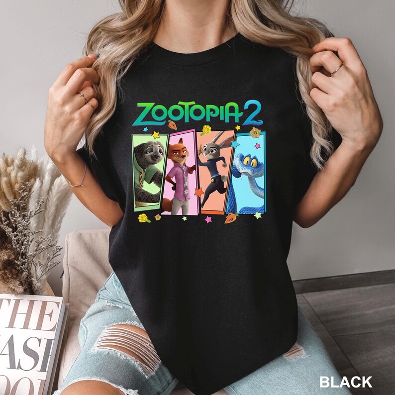 K&ouml;nnte beinhalten: Schwarzes T-Shirt mit dem Text "ZOOTOPIA 2" in T&uuml;rkis und einer bunten Grafik von Charakteren aus dem Animationsfilm. Die Grafik zeigt ein Faultier, einen Fuchs, ein Kaninchen und eine Schlange. Das Wort "BLACK" ist unten aufgedruckt.