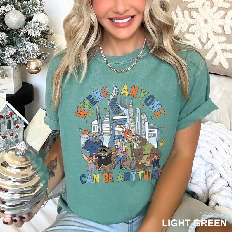 May include: Light green t-shirt featuring the text "WHERE ANYONE CAN BE ANYTHING" in a colourful, retro font. The graphic depicts cartoon characters and city buildings. The shirt is a short-sleeved, casual style.