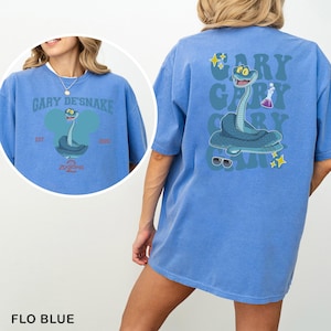 May include: Flo blue t-shirt featuring a cartoon snake design. The front of the shirt has the text "GARY DE'SNAKE" above a snake illustration. The back of the shirt repeats the text "GARY" with a snake illustration and sunglasses.