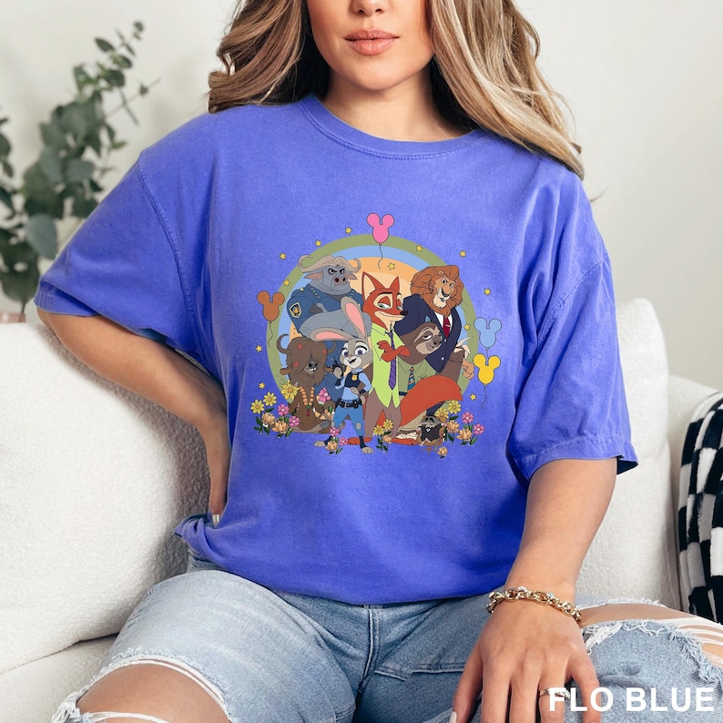 May include: A blue t-shirt featuring a colorful graphic of characters from the animated movie Zootopia. The design includes Judy Hopps, Nick Wilde, and other characters, surrounded by flowers and a rainbow. The text "FLO BLUE" is visible at the bottom.