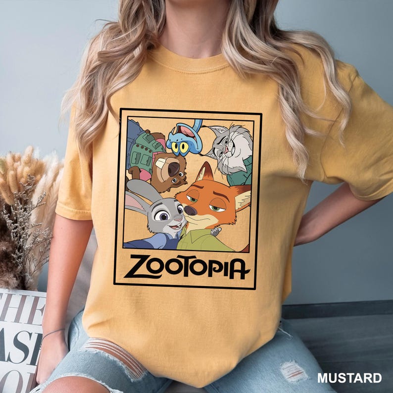 May include: Mustard-colored t-shirt featuring a framed graphic of characters from the animated film "Zootopia." The graphic includes Judy Hopps, Nick Wilde, and other characters. The word "Zootopia" is printed in a bold, black font below the image.