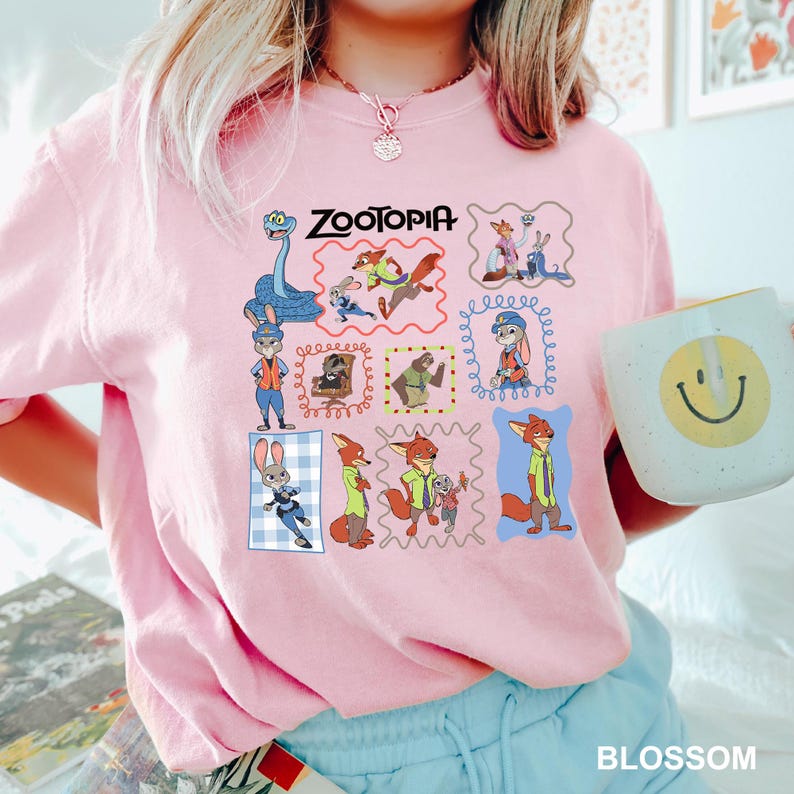 May include: Pink Zootopia-themed sweatshirt featuring a grid of character illustrations. The word "Zootopia" is printed across the top. A white mug with a yellow smiley face is held in the right hand. The word "BLOSSOM" is printed at the bottom.