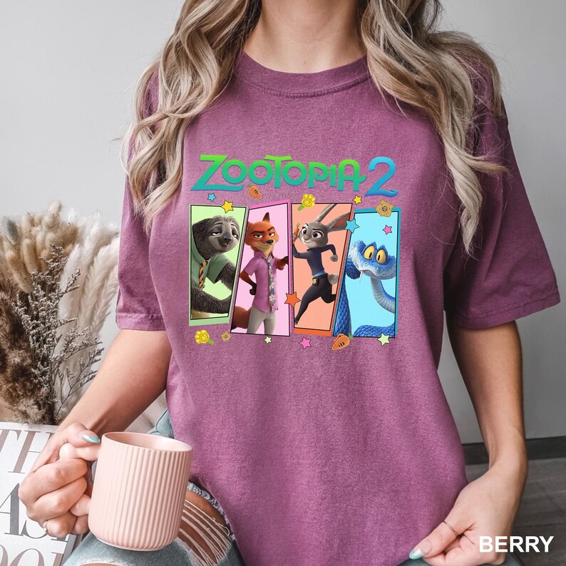 K&ouml;nnte beinhalten: Beerenfarbenes T-Shirt mit dem Text "ZOOTOPIA 2" und einer Grafik von Charakteren aus dem Film. Die Grafik zeigt ein Faultier, einen Fuchs, ein Kaninchen und eine Schlange in farbenfrohen Panels. Das Shirt ist ein l&auml;ssiger, bequemer Stil.