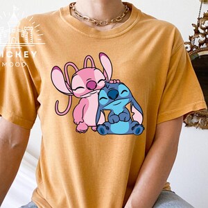 May include: A mustard yellow t-shirt with a graphic of Stitch and Angel from the Disney movie Lilo & Stitch. Stitch is blue and Angel is pink.
