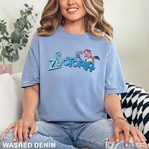 Op de afbeelding: Een lichtblauw T-shirt met korte mouwen met het woord "Zootopia" in een gestileerd lettertype, met stripfiguren. Het shirt is gemaakt van een zacht materiaal en is onderaan voorzien van het label "Washed Denim".
