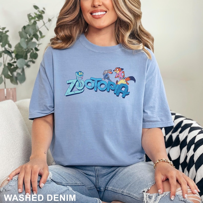 Op de afbeelding: Een lichtblauw T-shirt met korte mouwen met het woord "Zootopia" in een gestileerd lettertype, met stripfiguren. Het shirt is gemaakt van een zacht materiaal en is onderaan voorzien van het label "Washed Denim".