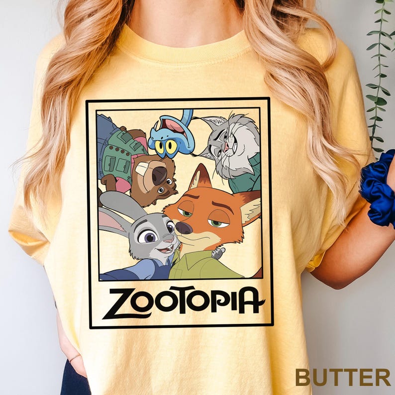 May include: Yellow t-shirt featuring a rectangular graphic of characters from the animated film Zootopia. The graphic includes Judy Hopps, Nick Wilde, and other characters. The word "Zootopia" is printed in black at the bottom of the graphic.