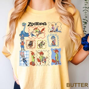 May include: Butter-colored t-shirt featuring a colorful graphic of characters from the animated film "Zootopia." The design includes various characters in framed illustrations, with the film's title at the top. The word "BUTTER" is printed at the bottom.