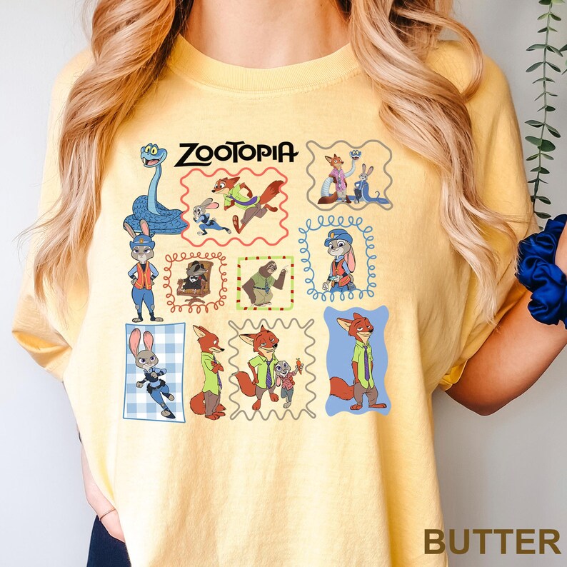 May include: Butter-colored t-shirt featuring a colorful graphic of characters from the animated film "Zootopia." The design includes various characters in framed illustrations, with the film's title at the top. The word "BUTTER" is printed at the bottom.