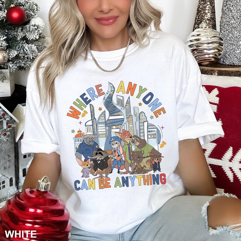 May include: White t-shirt with a colourful graphic featuring characters and a cityscape. The text "WHERE ANYONE CAN BE ANYTHING" is displayed in a rainbow font. The shirt is a casual style, suitable for everyday wear.