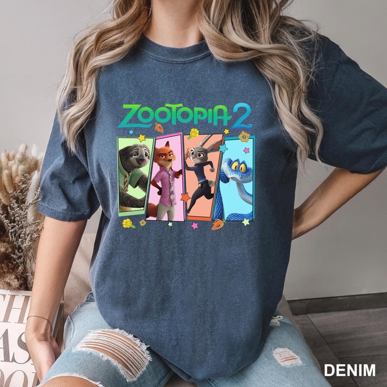 K&ouml;nnte beinhalten: Denimblaues T-Shirt mit dem Text "ZOOTOPIA 2" und einer Grafik von Zootopia-Charakteren. Die Grafik zeigt ein Faultier, einen Fuchs, ein Kaninchen und eine Schlange in farbenfrohen Panels. Das Shirt ist ein l&auml;ssiger Stil.