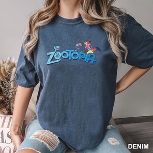 Op de afbeelding: Een donkerblauwe t-shirt met het woord "Zootopia" in blauw en stripfiguren. De t-shirt heeft korte mouwen en een ronde hals. Het woord "DENIM" staat rechtsonder in het wit gedrukt.