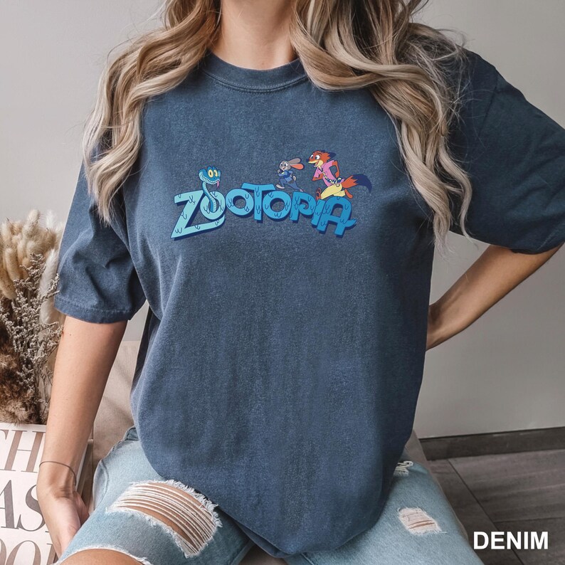 Op de afbeelding: Een donkerblauwe t-shirt met het woord "Zootopia" in blauw en stripfiguren. De t-shirt heeft korte mouwen en een ronde hals. Het woord "DENIM" staat rechtsonder in het wit gedrukt.